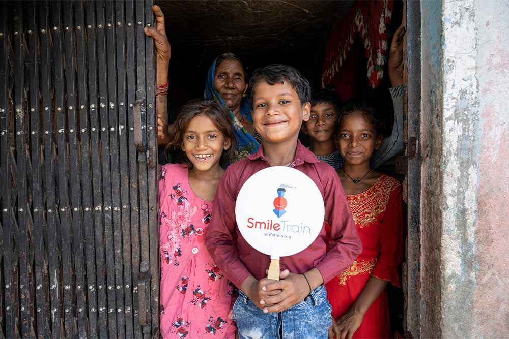 Shubham smiling and holding a Smile Train sign with his family after cleft surgery